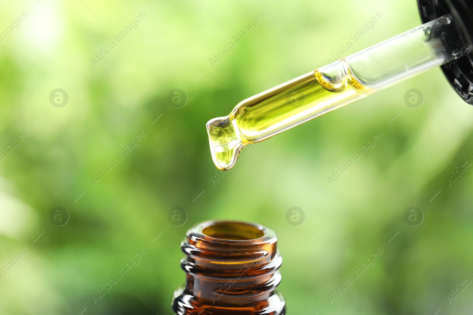 Photo of Dripping essential oil from pipette into glass bottle against blurred green background, closeup