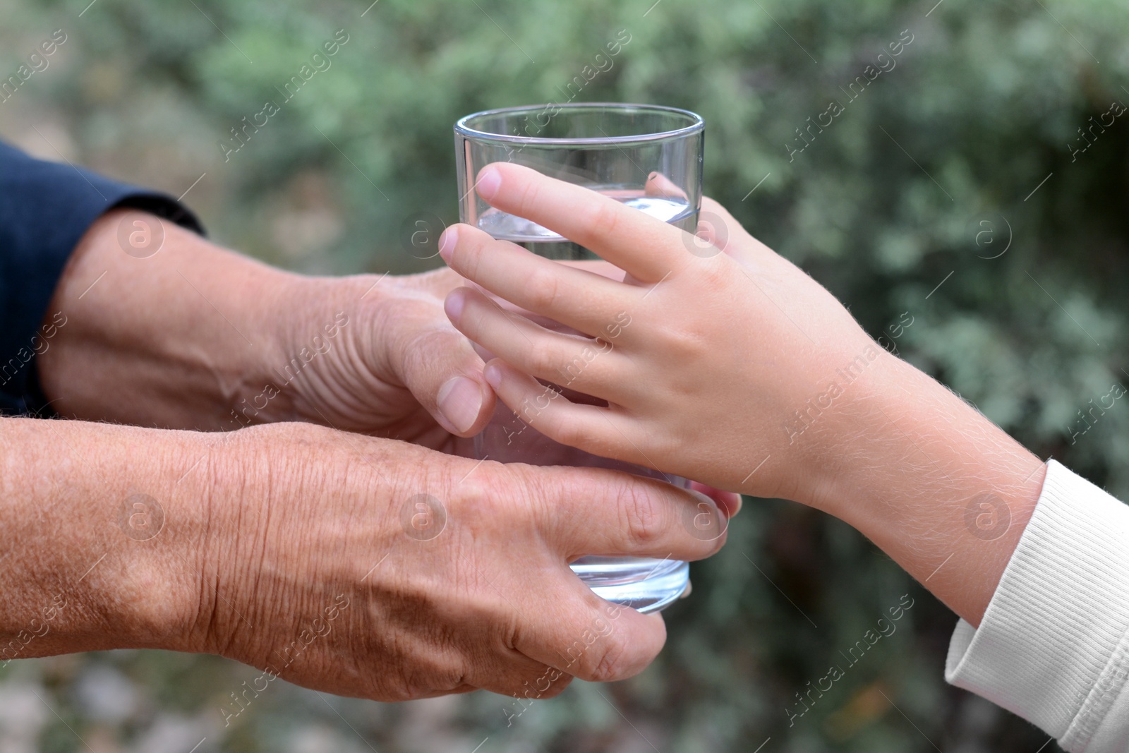 Photo of Child giving glass of water to elderly woman outdoors, closeup