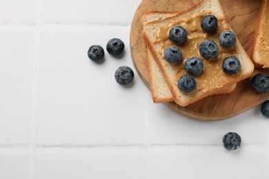 Photo of Delicious toasts with peanut butter and blueberries on white tiled table, top view. Space for text