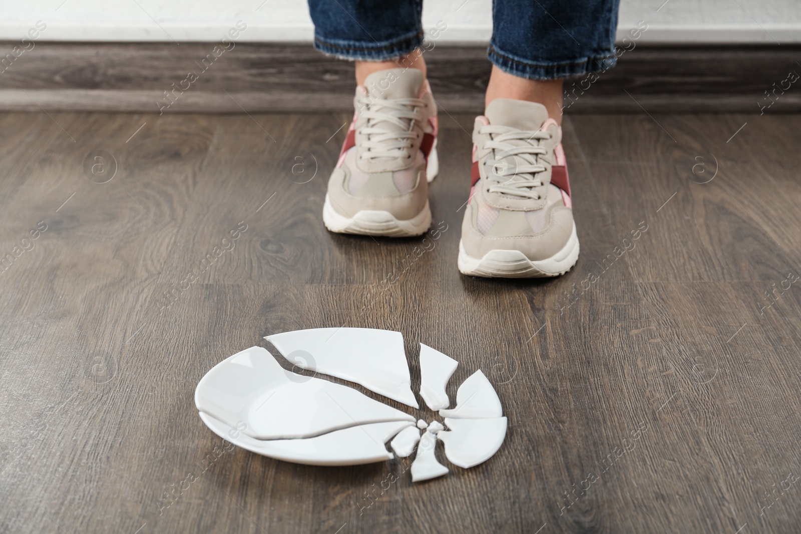 Photo of Woman in sneakers standing near broken plate on floor indoors, closeup