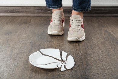 Photo of Woman in sneakers standing near broken plate on floor indoors, closeup