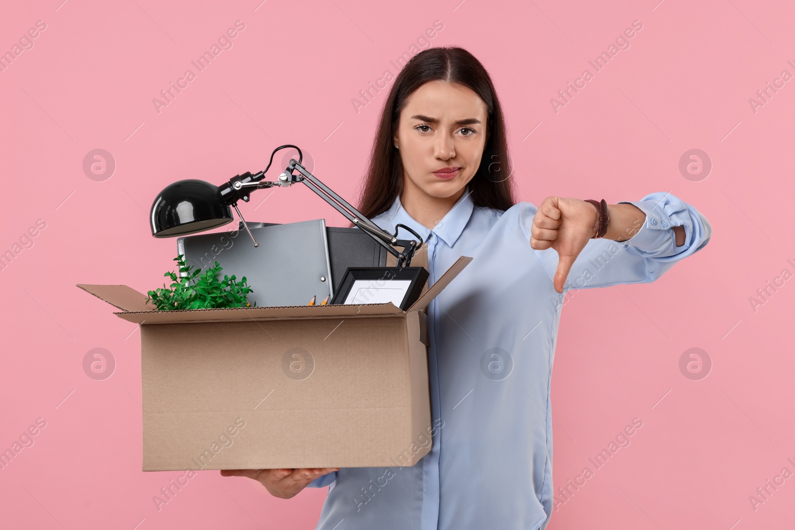 Photo of Unemployment problem. Unhappy woman with box of personal office belongings showing thumbs down on pink background