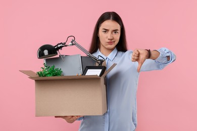 Unemployment problem. Unhappy woman with box of personal office belongings showing thumbs down on pink background