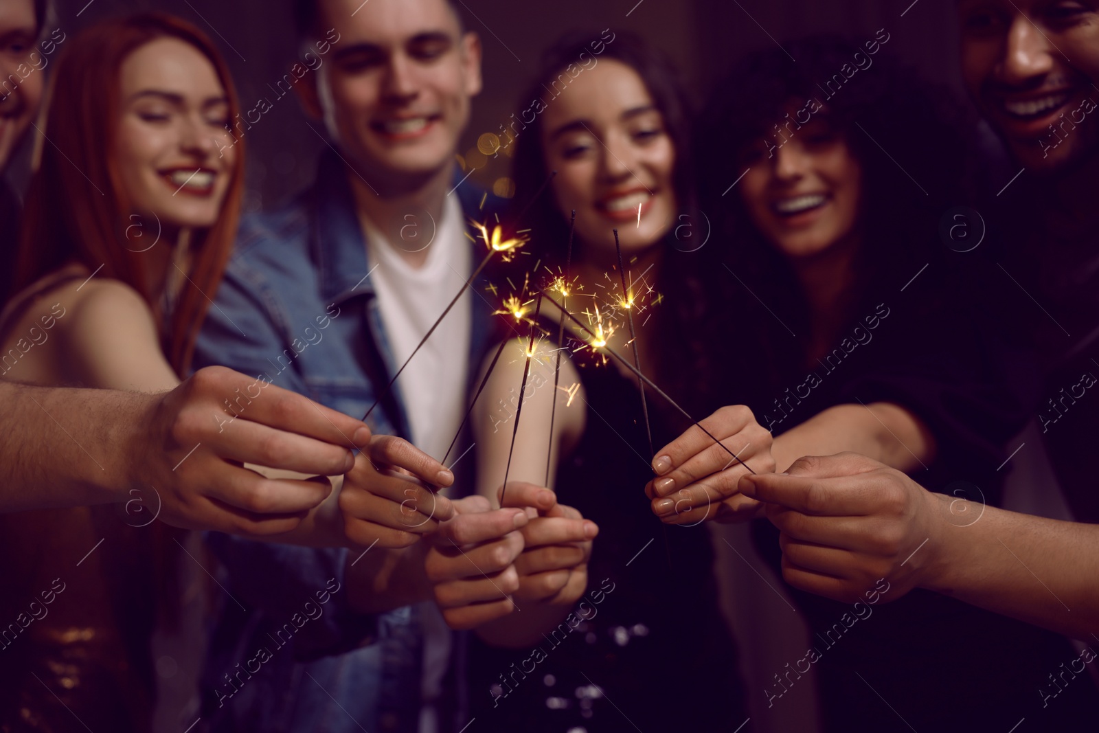 Photo of Happy friends with sparklers celebrating birthday indoors, focus on hands