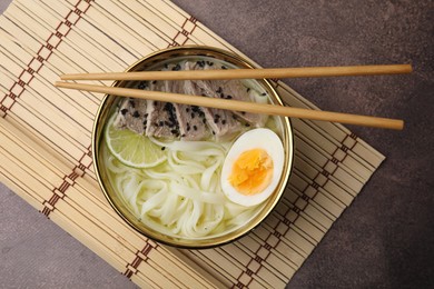 Photo of Bowl of delicious rice noodle soup with meat and egg on brown table, top view