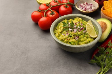Bowl of delicious guacamole, nachos chips and ingredients on grey table. Space for text