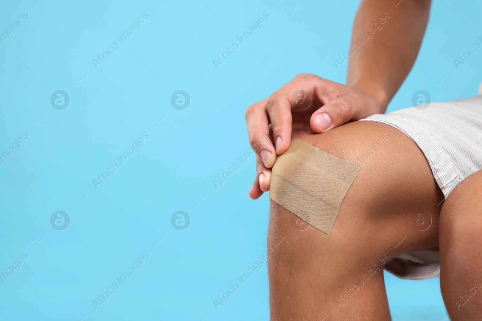 Photo of Man putting sticking plaster onto knee on light blue background, closeup. Space for text