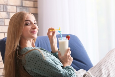 Young woman with glass of delicious milk shake in armchair indoors