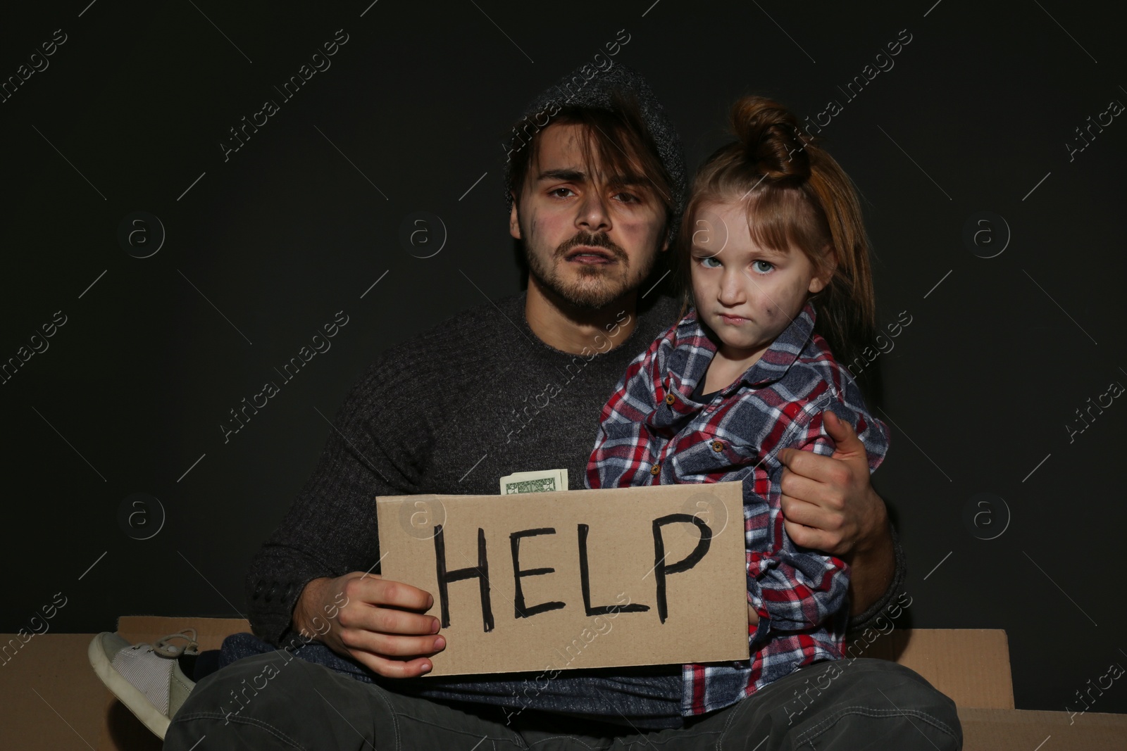 Photo of Poor father and daughter with HELP sign on floor near dark wall