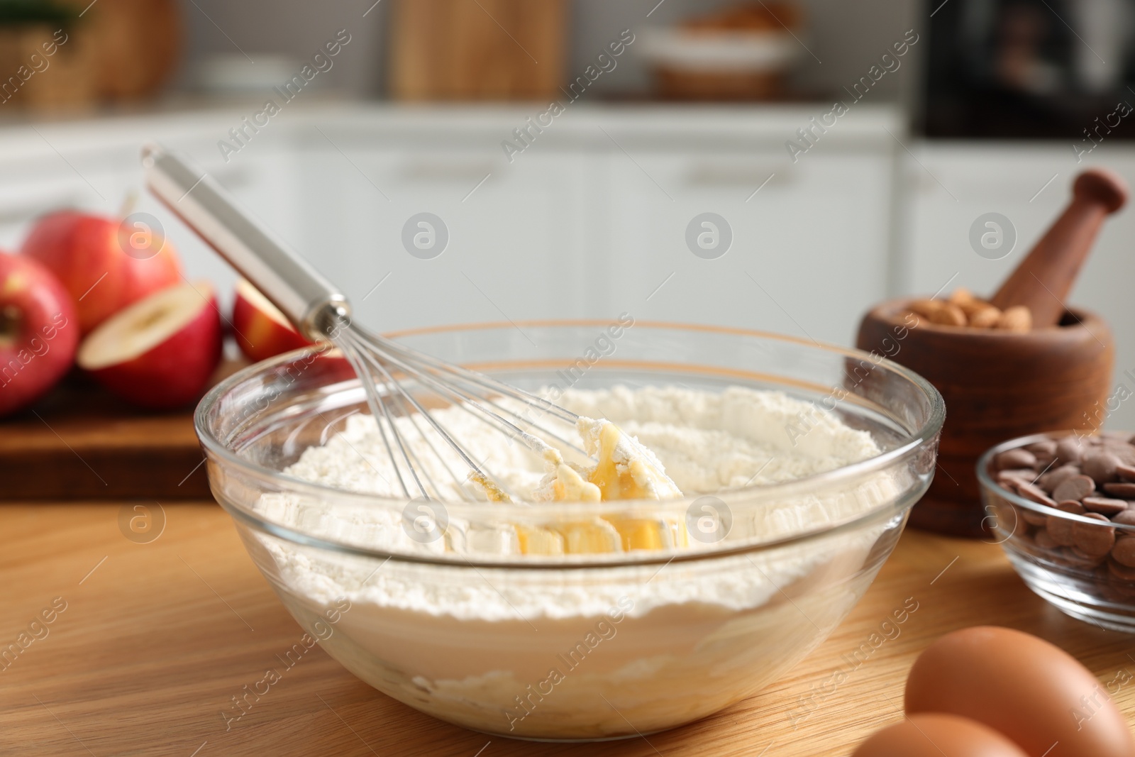 Photo of Cooking process. Metal whisk, bowl, flour, eggs and other products on wooden table in kitchen, closeup