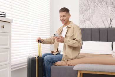 Photo of Smiling guest with suitcase and smartphone on bed in stylish hotel room