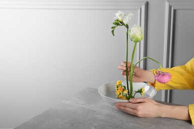 Photo of Stylish ikebana as house decor. Woman creating floral composition with fresh flowers at grey table near white wall, closeup and space for text