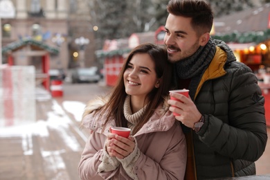 Photo of Young couple with cups of mulled wine at winter fair. Space for text