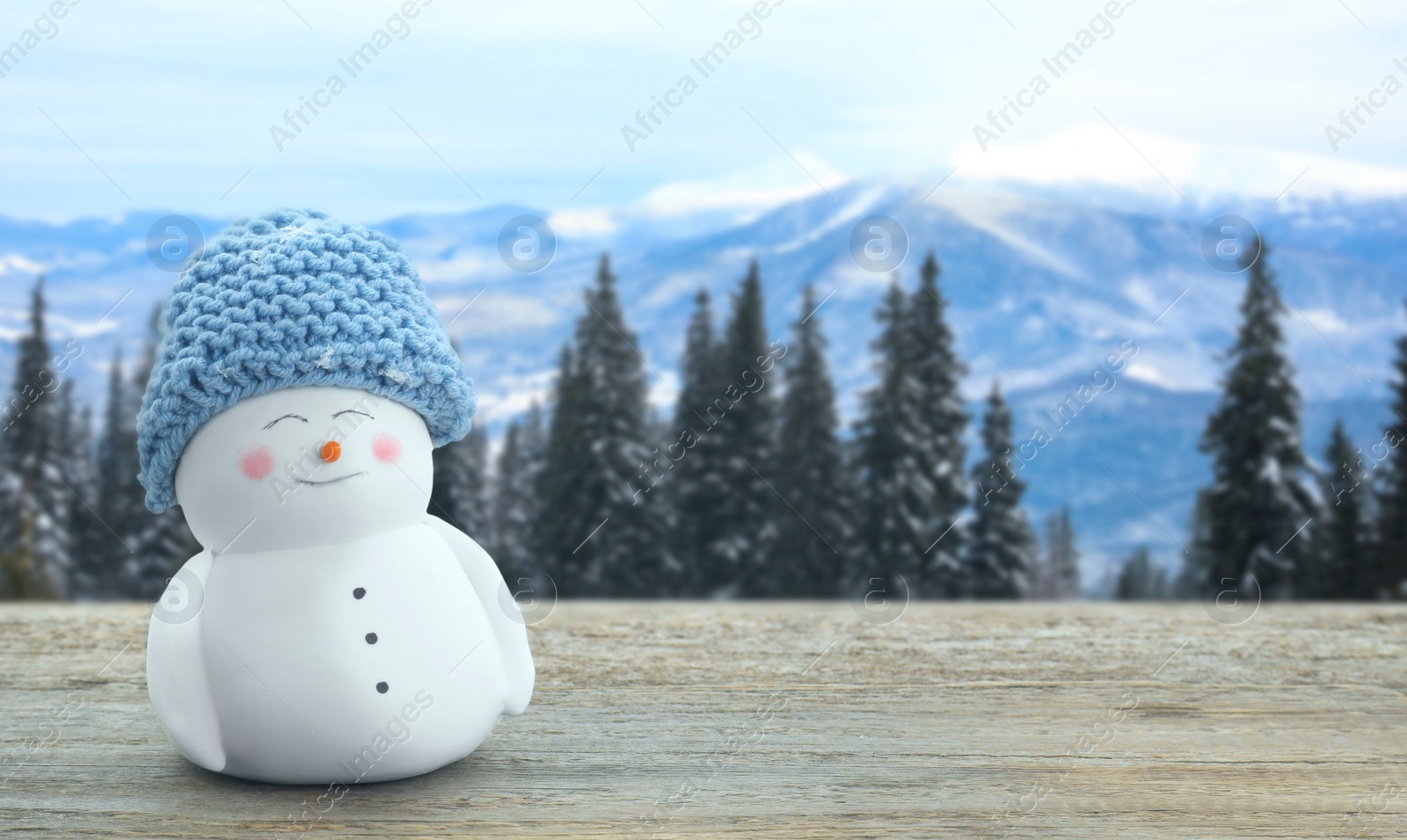 Image of Cute decorative snowman in blue hat on wooden table in mountains, space for text. Banner design