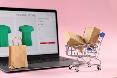 Photo of Online store. Laptop, mini shopping cart and purchases on pink background