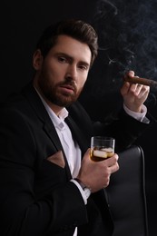 Photo of Handsome man in elegant suit with glass of whiskey smoking cigar on black background