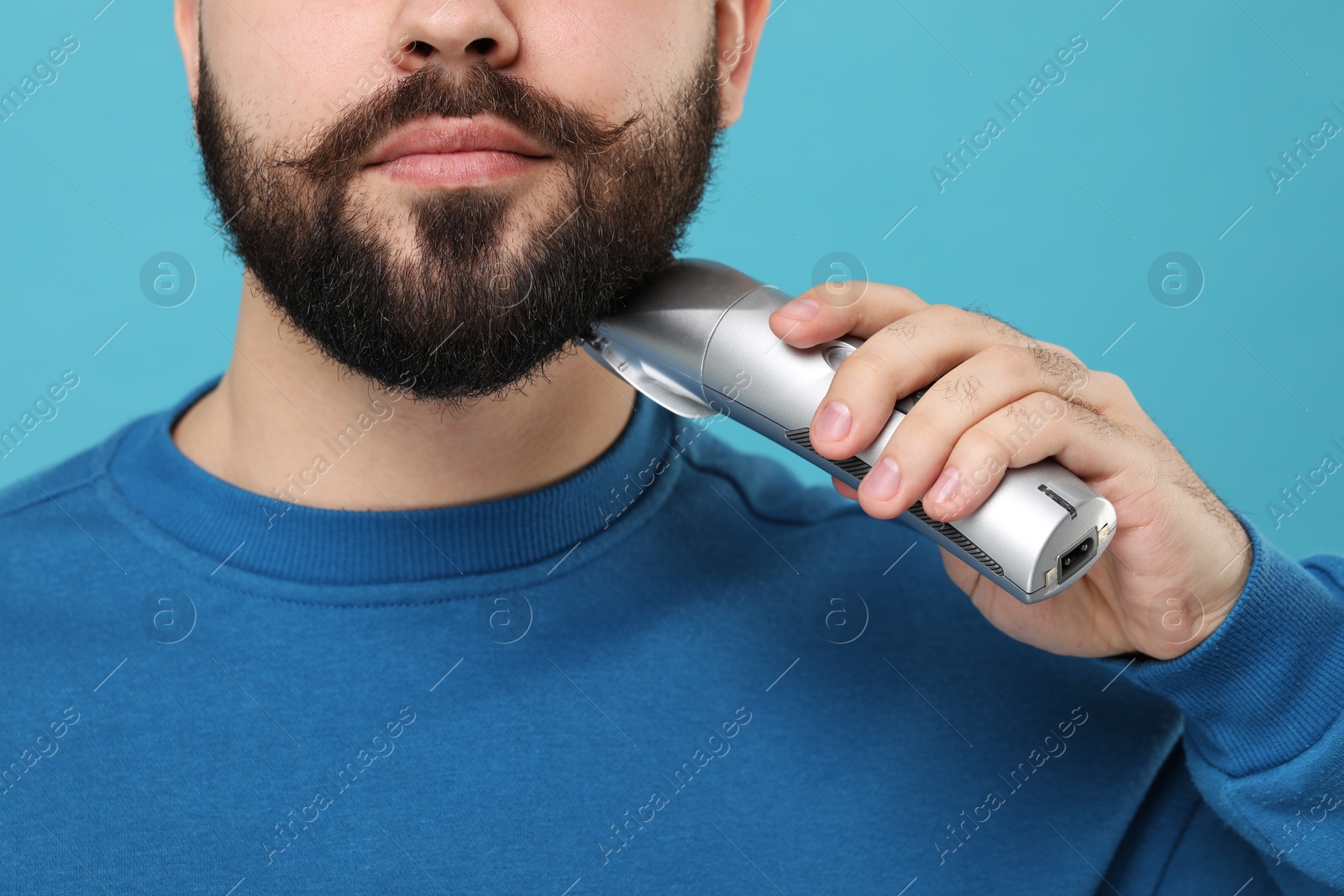 Photo of Handsome young man trimming beard on light blue background, closeup