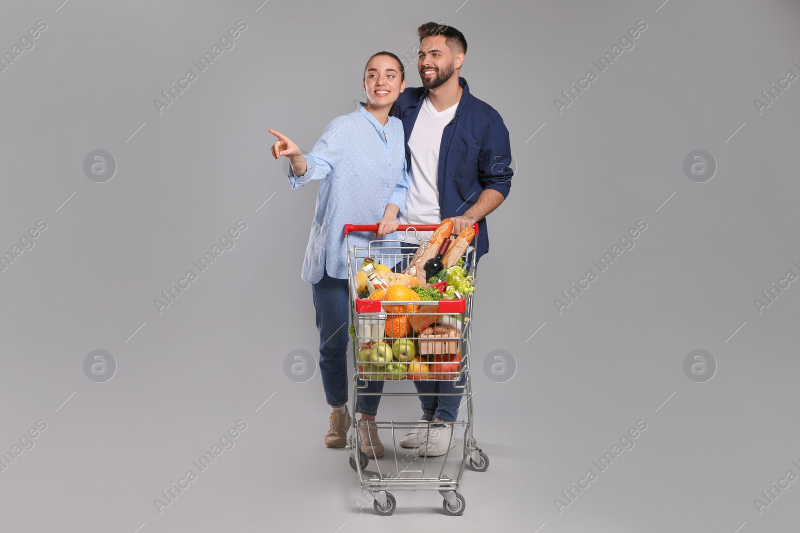 Photo of Happy couple with shopping cart full of groceries on light grey background