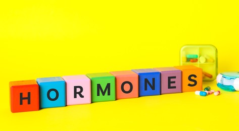 Photo of Word Hormones made of colorful cubes with letters and pills on yellow background