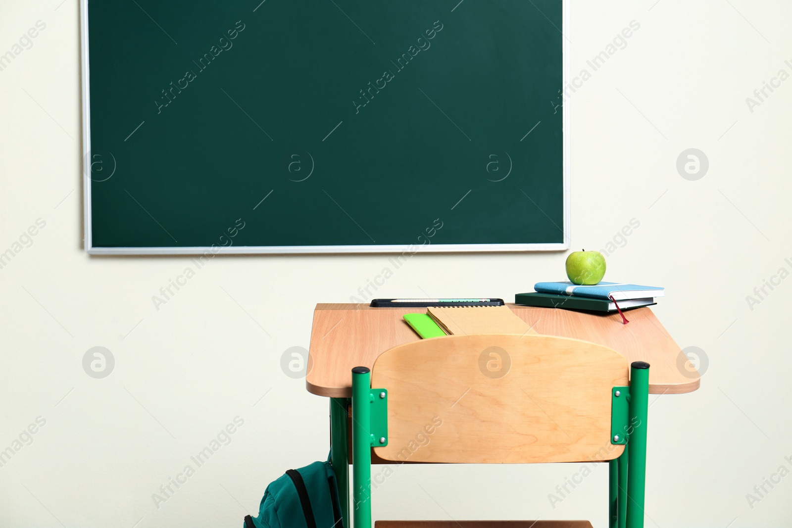 Photo of Wooden school desk with stationery, apple and backpack near chalkboard in classroom