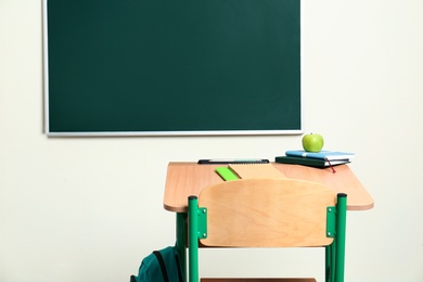 Wooden school desk with stationery, apple and backpack near chalkboard in classroom