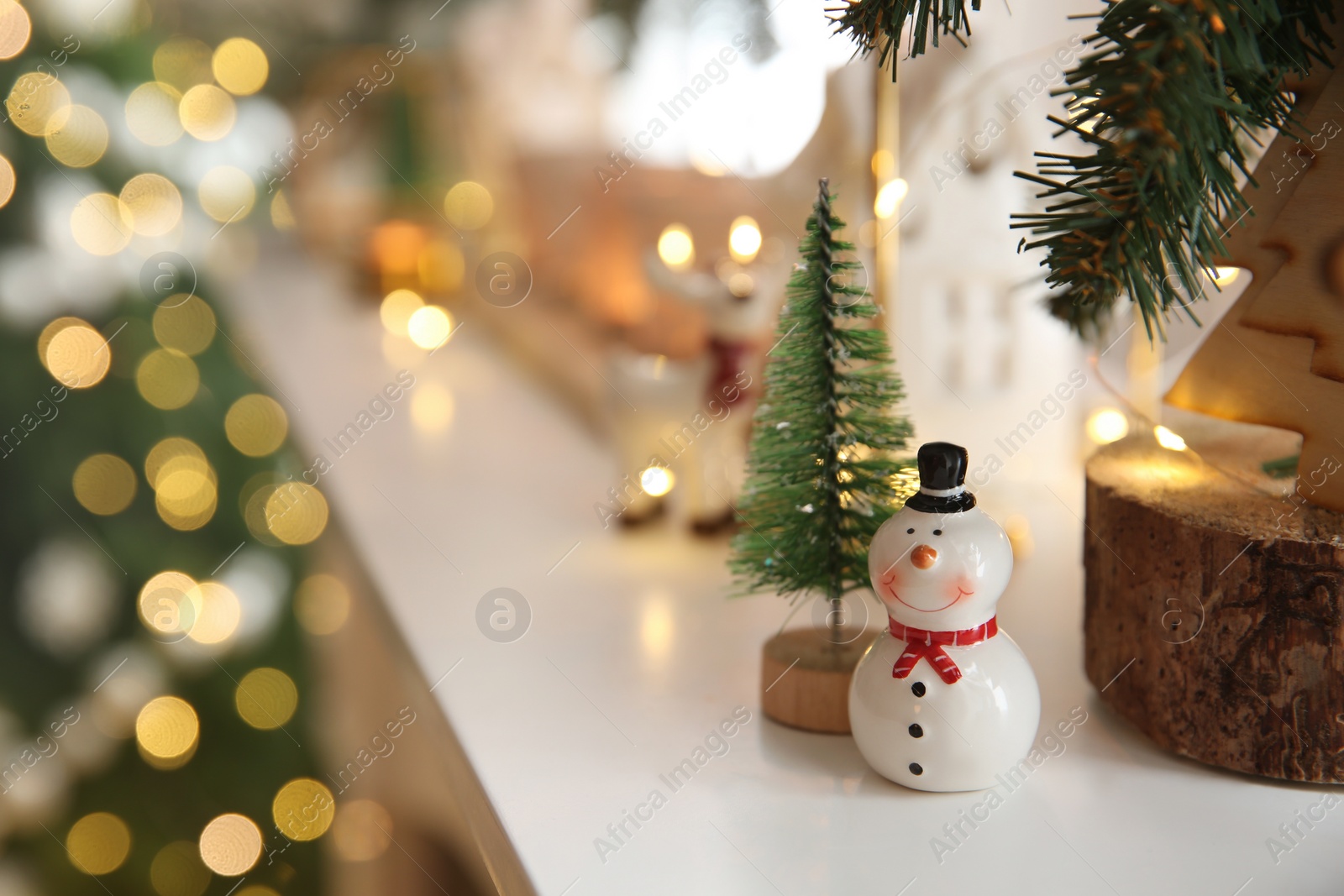 Photo of Funny ceramic snowman and small decorative tree on white table against blurred lights. Space for text