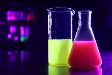 Photo of Laboratory glassware with luminous liquids on table against dark background, space for text