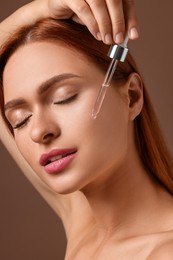 Beautiful young woman applying cosmetic serum onto her face on brown background, closeup