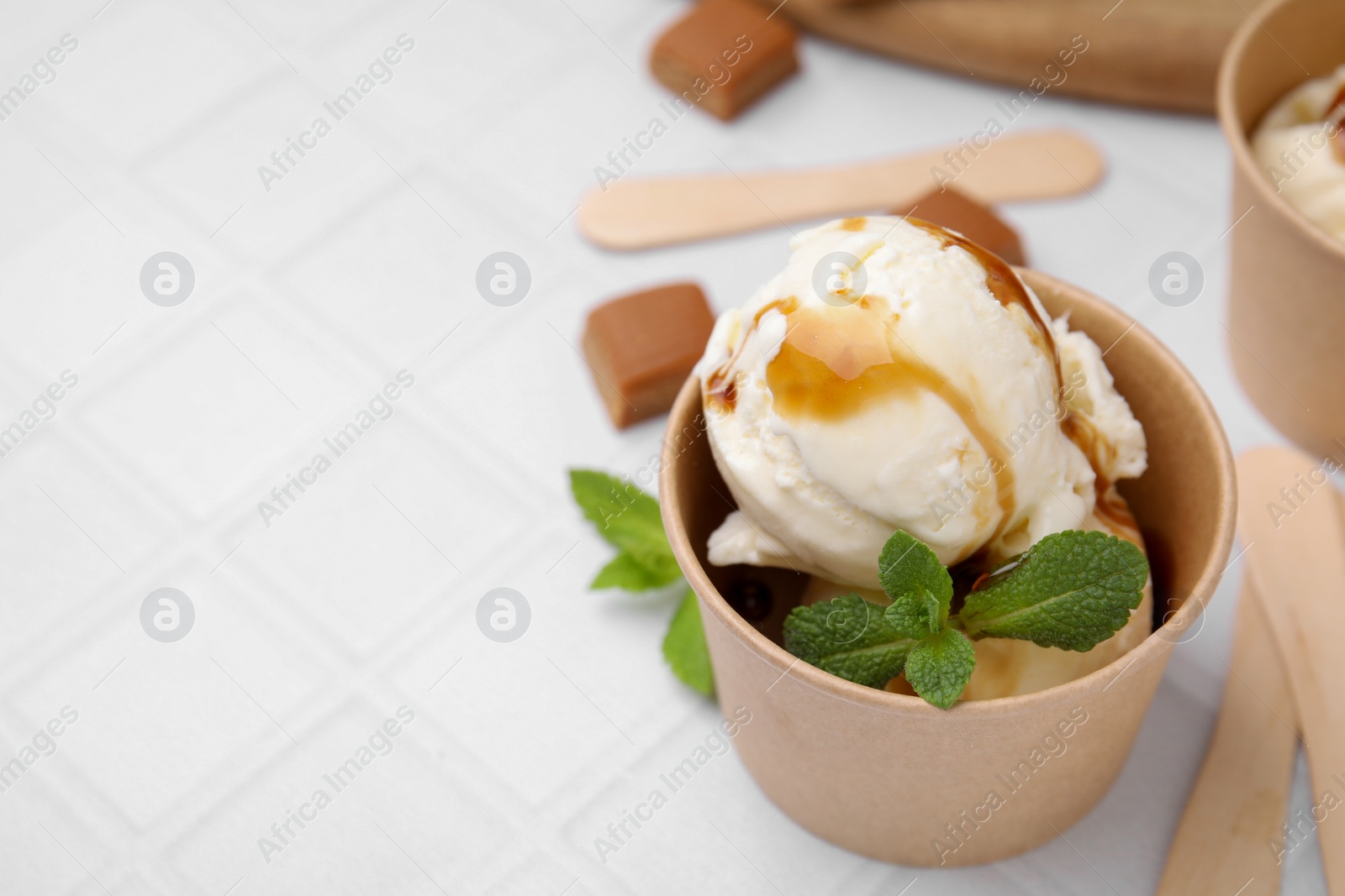 Photo of Scoops of ice cream with caramel sauce, mint leaves and candies on white tiled table, closeup. Space for text