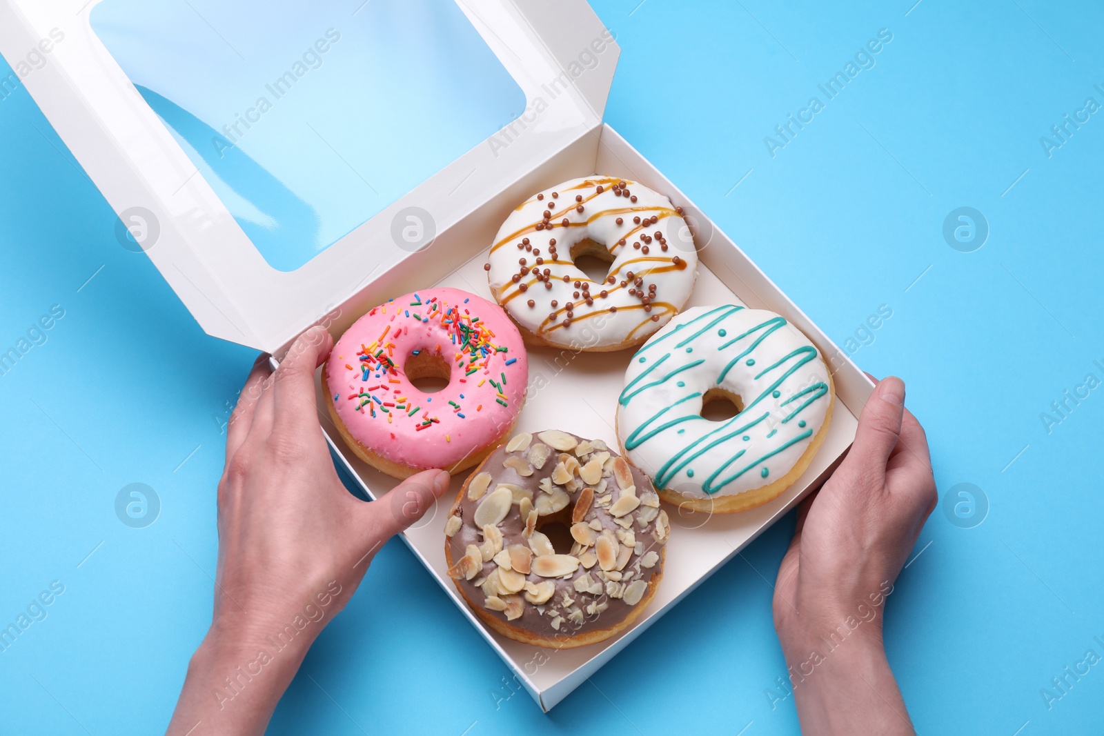 Photo of Woman taking tasty glazed donut from box on light blue background, top view