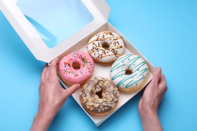 Woman taking tasty glazed donut from box on light blue background, top view