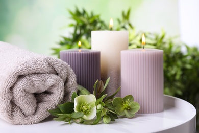 Photo of Burning candles, towel and flowers on white table against blurred green background, space for text