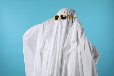 Photo of Stylish ghost. Person covered with white sheet in sunglasses and headphones on light blue background