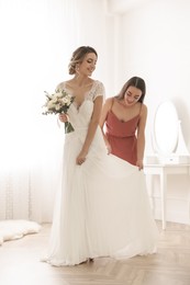 Young woman helping bride to put on wedding dress in room