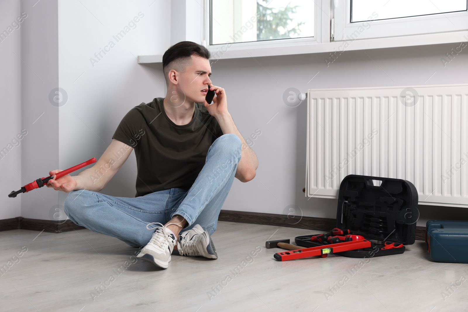 Photo of Handyman with pipe wrench talking on phone near radiator in room