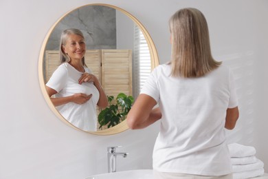 Beautiful senior woman doing breast self-examination near mirror in bathroom