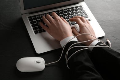 Photo of Internet addiction. Closeup of man typing on laptop at black table, hands tied with computer mouse cable