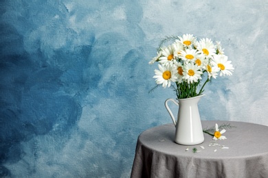 Photo of Jug with beautiful chamomile flowers on table against color background