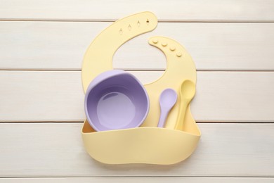 Photo of Silicone baby bib and plastic dishware on white wooden background, top view