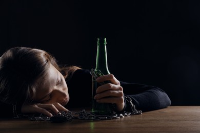 Photo of Alcohol addiction. Woman chained with bottle of beer sleeping at wooden table against black background