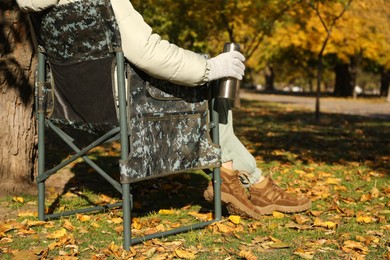 Woman with thermos sitting in camping chair outdoors on autumn sunny day, closeup