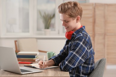Photo of Online learning. Smiling teenage boy writing in notebook near laptop at home