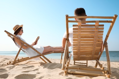 Photo of Young woman reading book and man relaxing in deck chairs on beach near sea