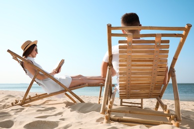 Young woman reading book and man relaxing in deck chairs on beach near sea