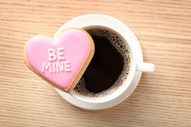 Photo of Heart shaped cookie with written phrase Be Mine and cup of coffee on wooden background, top view. Valentine's day treat