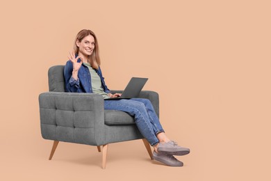 Photo of Happy woman with laptop sitting in armchair and showing ok gesture on beige background
