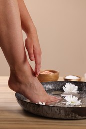 Photo of Woman soaking her foot in bowl with water, spa stones and chrysanthemum flowers on wooden surface, closeup. Pedicure procedure