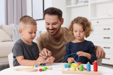 Photo of Motor skills development. Father and his kids playing with wooden pieces and string for threading activity at table indoors
