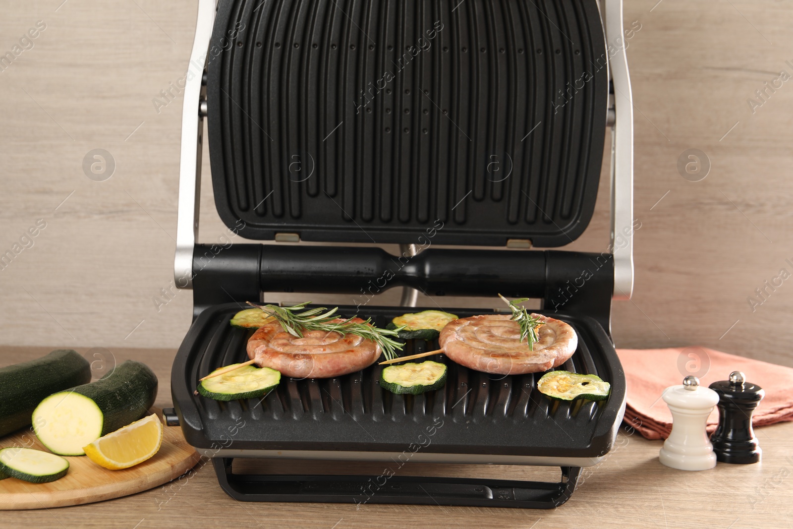 Photo of Electric grill with homemade sausages, zucchini and rosemary on wooden table