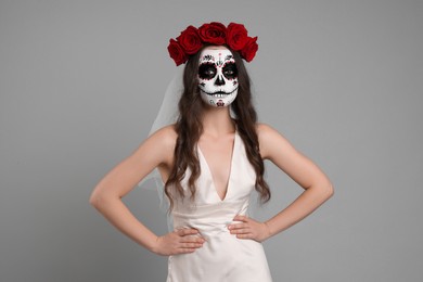 Young woman in scary bride costume with sugar skull makeup and flower crown posing on light grey background. Halloween celebration